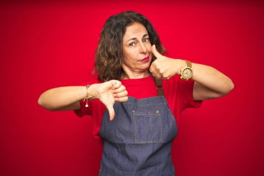 Orta yaş yaşlı kadın kırmızı izole arka plan üzerinde önlük üniforma giyen başparmak yukarı ve aşağı, anlaşmazlık ve anlaşma ifade yapıyor. Çılgın çatışma