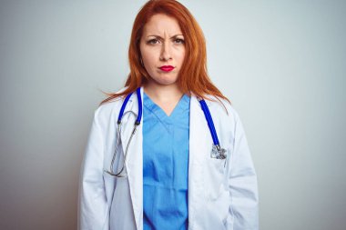 Genç kızıl saçlı doktor kadın beyaz izole arka plan şüpheci ve sinir üzerinde stetoskop kullanarak, sorun nedeniyle üzgün kaşlarını çatma. Negatif kişi.
