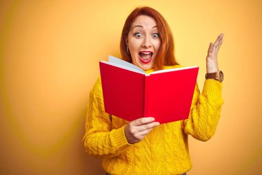 Genç kızıl saçlı öğretmen kadın sarı izole arka plan üzerinde kırmızı kitap okuma çok mutlu ve heyecanlı, kazanan ifade büyük bir gülümseme ile çığlık zafer kutluyor ve ellerini kaldırdı