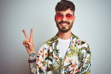 Tatilde yazlık gömlek güneş gözlüğü takan dövmeli adam izole beyaz arka plan üzerinde mutlu yüz ile gülümseyerek zafer işareti yapıyor kameraya göz kırparak. İki numara.