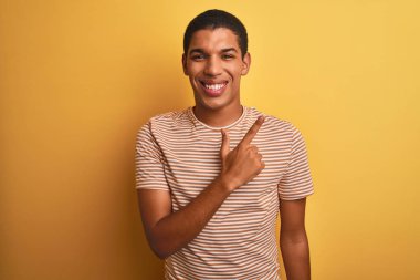 Genç yakışıklı arap adam çizgili t-shirt giyen izole sarı arka plan neşeli bir yüz yüzü ile işaret eden bir gülümseme ile neşeli yüzünde mutlu ve doğal ifade ile yan yana