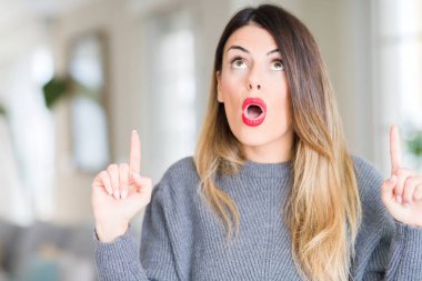 Evde kış kazak giyen genç güzel kadın hayrete ve yukarıya bakıyor ve parmakları ile işaret ve silah kaldırdı şaşırttı.