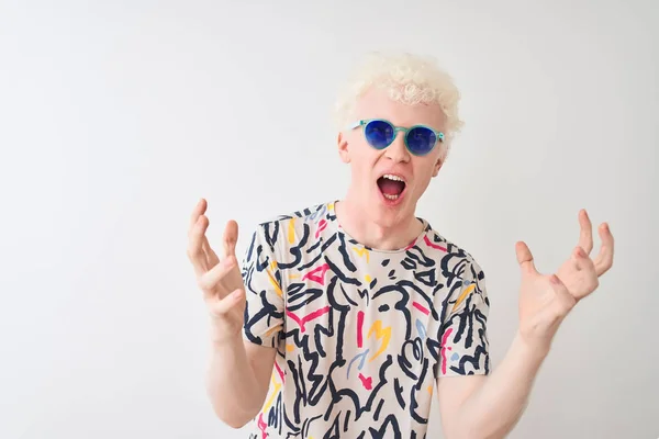 Young albino blond man wearing colorful t-shirt and sunglasses over isolated red background crazy and mad shouting and yelling with aggressive expression and arms raised. Frustration concept.