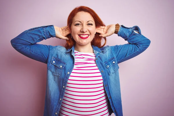 Beautiful redhead woman wearing denim shirt and striped t-shirt over isolated pink background Smiling pulling ears with fingers, funny gesture. Audition problem