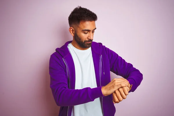 Young indian man wearing purple sweatshirt standing over isolated pink background Checking the time on wrist watch, relaxed and confident