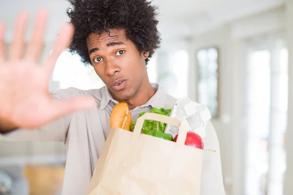 African American man holding groceries bag with fresh vegetables at home with open hand doing stop sign with serious and confident expression, defense gesture