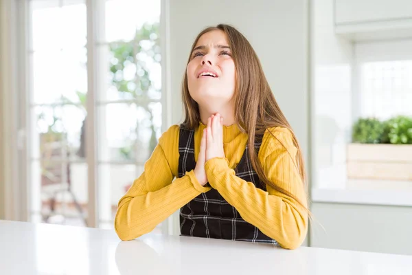 Young beautiful blonde kid girl wearing casual yellow sweater at home begging and praying with hands together with hope expression on face very emotional and worried. Asking for forgiveness. Religion concept.