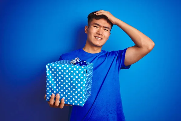 Young asian chinese man holding birthday gift standing over isolated blue background stressed with hand on head, shocked with shame and surprise face, angry and frustrated. Fear and upset for mistake.