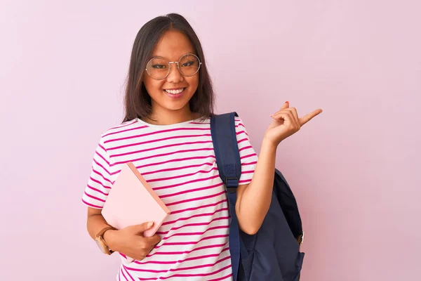 Chinese student woman wearing glasses backpack holding book over isolated pink background very happy pointing with hand and finger to the side
