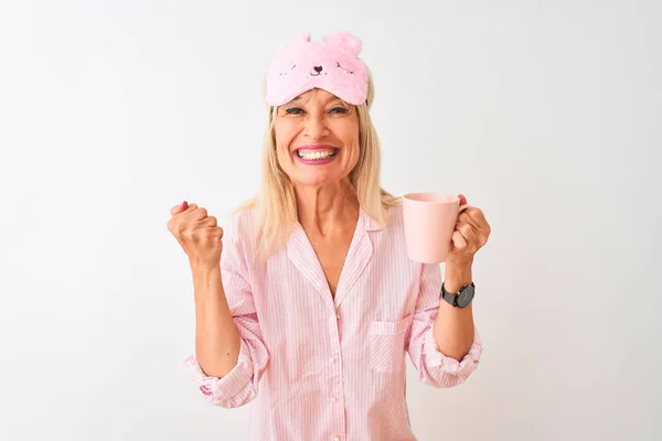 Middle age woman wearing sleep mask pajama drinking coffee over isolated white background screaming proud and celebrating victory and success very excited, cheering emotion