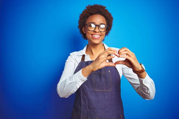 Young african american woman shop owner wearing business apron over blue background smiling in love showing heart symbol and shape with hands. Romantic concept.