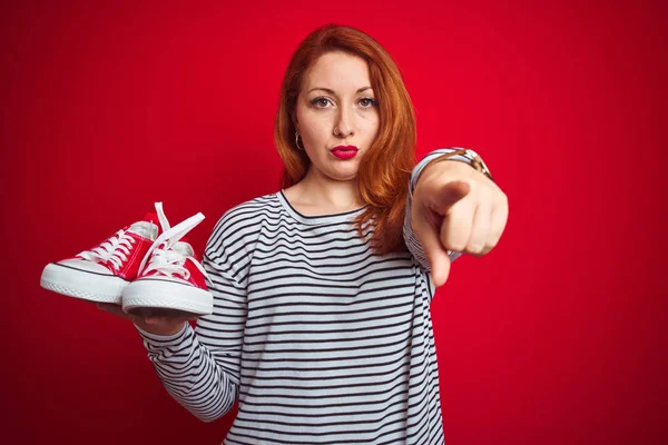 年轻美丽的红发女人拿着运动鞋在红色孤立的背景 用手指指着相机和你 手标志 积极和自信的姿态从前面 — 图库照片