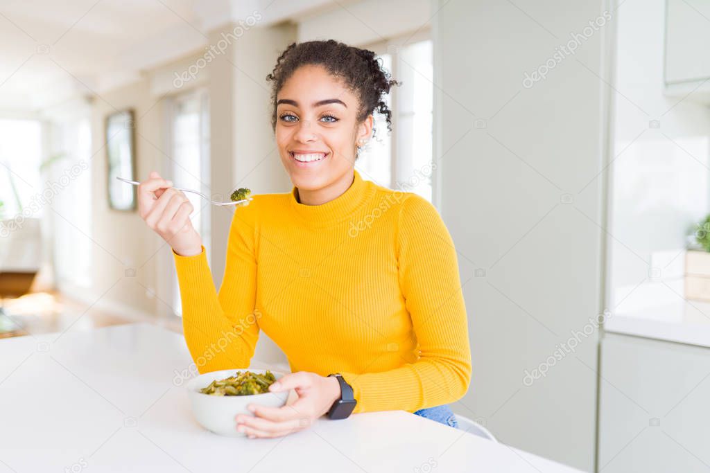Young african american woman eating healthy green vegatables with a happy face standing and smiling with a confident smile showing teeth