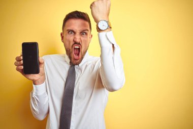 Genç yakışıklı iş adamı sarı izole arka plan üzerinde akıllı telefon ekranı gösteren kızgın ve sinirli öfke ile bağırarak, deli ve yükseltilmiş el ile bağırıyor, öfke kavramı