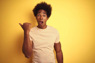 Afro saçlı, çizgili tişört giyen Amerikalı adam izole sarı arka plan üzerinde duran yan el parmağı ile işaret sürpriz, açık ağız hayrete ifade.