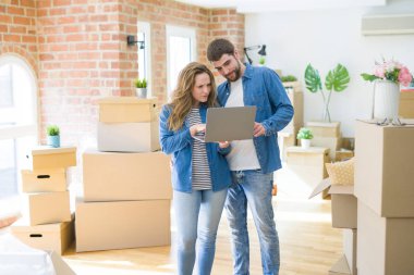 Yeni bir daireye taşımak için mutlu karton kutular etrafında bir odada bilgisayar laptop ayakta kullanarak genç çift