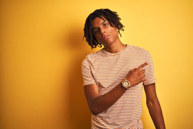 Çizgili t-shirt giyen dreadlocks giyen Afro adam izole sarı arka plan üzerinde duran yan reklam gösteren el parmağı ile işaret, ciddi ve sakin yüz