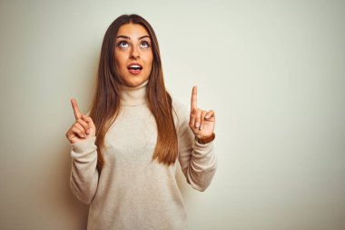 Izole beyaz arka plan üzerinde kış boğazlı kazak giyen genç güzel kadın şaşırttı ve yukarı bakarak ve parmakları ile işaret ve kolları kaldırdı şaşırttı.