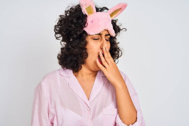 Izole beyaz arka plan üzerinde pijama ve uyku maskesi giyen kıvırcık saçlı Arap kadın el ile ağız kapsayan yorgun esneme sıkılmış. Huzursuz ve uykululuk.