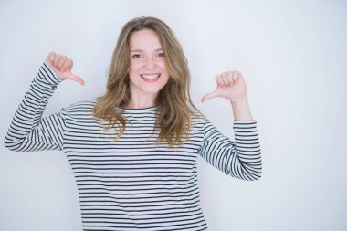 Beyaz izole arka plan üzerinde çizgili kazak giyen mavi gözlü güzel sarışın kız yüzünde gülümseme ile emin görünümlü, parmakları gururlu ve mutlu kendini işaret.