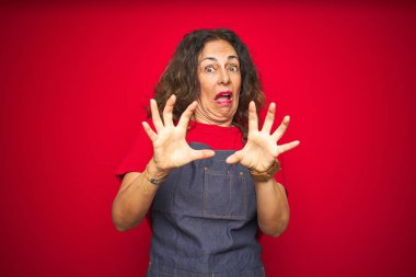 Kırmızı izole arka plan üzerinde önlük üniforma giyen orta yaş yaşlı kadın korkmuş ve korku ifade ile korkmuş elleriyle jest durdurmak, şok içinde bağırarak. Panik kavramı.
