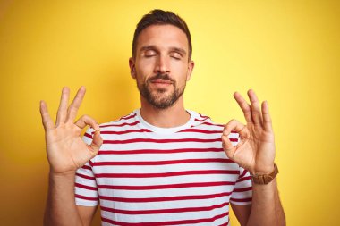 Genç yakışıklı adam sarı izole arka plan üzerinde rahat kırmızı çizgili t-shirt giyen rahat ve gözleri parmakları ile meditasyon jest yapıyor kapalı gülümseyerek. Yoga konsepti.