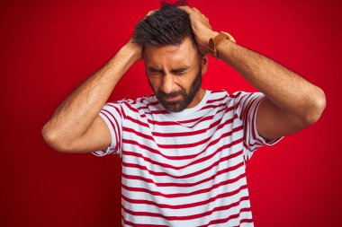 Çizgili t-shirt giyen genç hintli adam izole kırmızı arka plan üzerinde baş ağrısı umutsuz acı ve stresli çünkü ağrı ve migren. Eller baş üstünde.