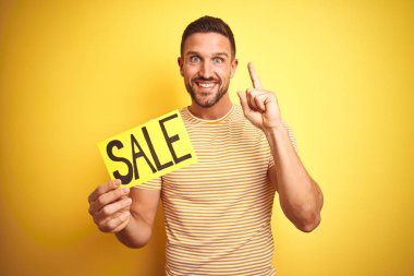 Sarı izole arka plan üzerinde satış reklam afiş idamı tutan genç adam mutlu yüz, bir numara ile bir fikir veya soru işaret parmağı ile şaşırttı