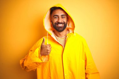 Genç hintli adam kapüşonlu izole sarı arka plan üzerinde duran el ile jest kadar mutlu başparmak yapıyor. Başarı göstererek kameraya bakan ifadeyi onaylama.