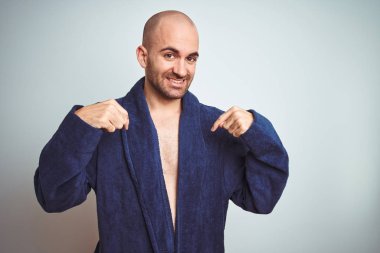Mavi bornoz giyen genç adam, izole arka plan üzerinde rahat bir yaşam tarzı yüzünde gülümseme ile emin görünümlü, parmakları gururlu ve mutlu kendini işaret.