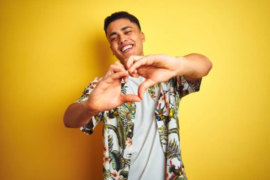 Genç Brezilyalı adam tatilde izole sarı arka plan üzerinde yazlık çiçek gömlek giyen aşk elleri ile kalp sembolü şekli yapıyor gülümseyerek. Romantik kavram.