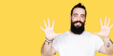 Uzun saç ve sakal gösterilen ve parmakları ile işaret casual beyaz t-shirt giyen genç hipster adamla kendinden emin ve mutlu gülümseyerek süre on numara.