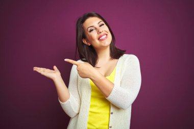Genç güzel esmer kadın mor izole arka plan üzerinde bir ceket şaşırttı ve el ile sunarken kameraya gülümseyerek ve parmak ile işaret.