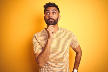 İzole sarı arka plan üzerinde duran tişört giyen genç hintli adam çenesi üzerinde soru, dalgın ifade yi düşünüyor. Düşünceli bir yüzle gülümsüyor. Şüphe kavramı.