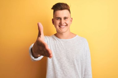 Genç yakışıklı adam beyaz rahat t-shirt izole sarı arka plan üzerinde ayakta giyen tebrik ve karşılama olarak dostça sunan el sıkışma gülümseyerek. Başarılı iş.