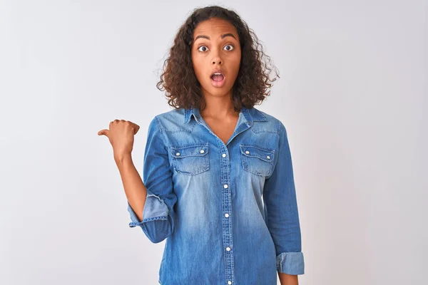 Young brazilian woman wearing denim shirt standing over isolated white background Surprised pointing with hand finger to the side, open mouth amazed expression.