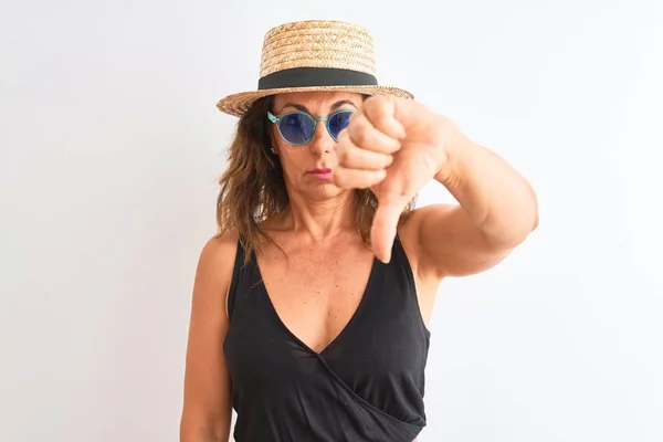 Middle age woman wearing black t-shirt sunglasses and hat over isolated white background with angry face, negative sign showing dislike with thumbs down, rejection concept