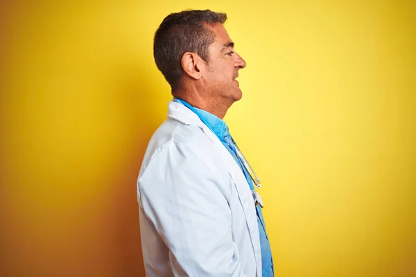 Handsome middle age doctor man wearing stethoscope over isolated yellow background looking to side, relax profile pose with natural face with confident smile.