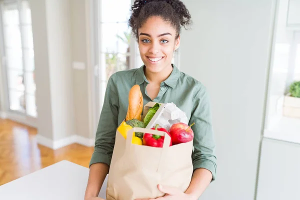 Young african american girl holding paper bag of groceries from supermarket with a happy face standing and smiling with a confident smile showing teeth