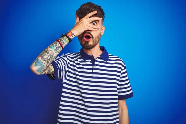 Çizgili polo giyen dövmeli genç adam izole mavi arka plan üzerinde duran şok yüz ve el ile gözleri kapsayan gözetleme, utanmış ifade ile parmaklarından bakarak.