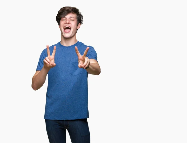 勝利のサインをやって指を表示するカメラを笑みを浮かべて孤立の背景に青い シャツを着ての若いハンサムな男 — ストック写真