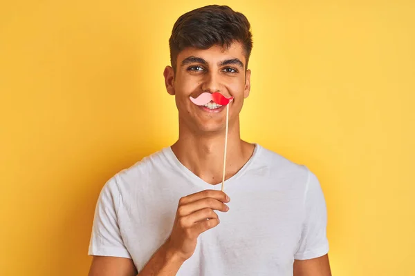 Young indian man holding fanny mustache standing over isolated yellow background with a happy face standing and smiling with a confident smile showing teeth