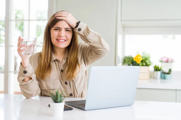 Beautiful young woman working with computer takes a break to drink glass of water stressed with hand on head, shocked with shame and surprise face, angry and frustrated. Fear and upset for mistake.