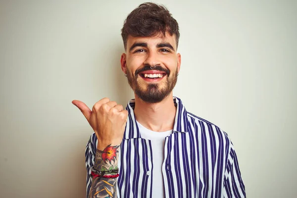 Young man with tattoo wearing striped shirt standing over isolated white background smiling with happy face looking and pointing to the side with thumb up.