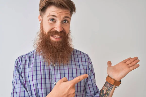 Redhead İrlandalı adam izole beyaz arka plan üzerinde kablosuz kulaklık kullanarak müzik dinlerken şaşırttı ve el ile sunarken ve parmak ile işaret ederken kameraya gülümseyerek.