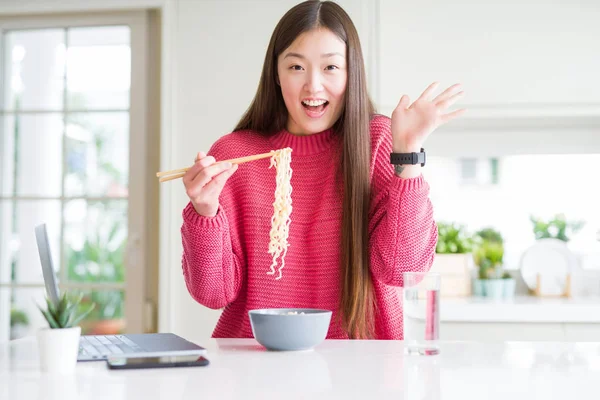 Güzel Asyalı kadın dizüstü bilgisayar kullanarak çalışan ve asya erişte yeme çok mutlu ve heyecanlı, kazanan ifade büyük bir gülümseme ile çığlık zafer kutluyor ve ellerini kaldırdı