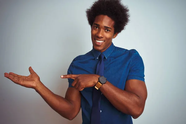 American business man with afro hair wearing blue shirt and tie over isolated white background amazed and smiling to the camera while presenting with hand and pointing with finger.