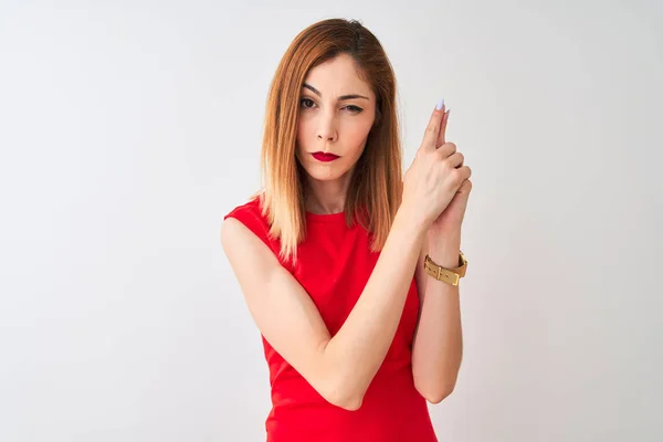 Redhead businesswoman wearing elegant red dress standing over isolated white background Holding symbolic gun with hand gesture, playing killing shooting weapons, angry face