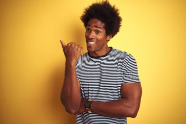Afro saçlı Afro-Amerikan adam izole sarı arka plan üzerinde lacivert çizgili t-shirt giyen mutlu yüz görünümlü ve başparmak ile yan işaret gülümseyerek.