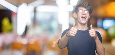 El ile pozitif jest yapıyor onaylama izole arka plan üzerinde gözlük takan genç yakışıklı adam başparmak gülümseyen ve başarı için mutlu. Kamera, kazanan jest arıyorsunuz.